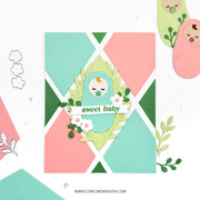 A handmade baby shower card featuring a pastel geometric background of pink, mint green, and dark green triangles. The center is decorated with a layered paper wreath, a sleeping baby illustration, and a banner that reads 'sweet baby'.