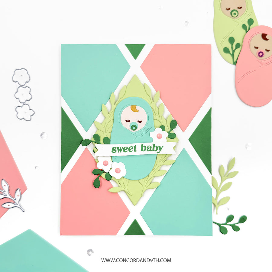 A handmade baby shower card featuring a pastel geometric background of pink, mint green, and dark green triangles. The center is decorated with a layered paper wreath, a sleeping baby illustration, and a banner that reads 'sweet baby'.