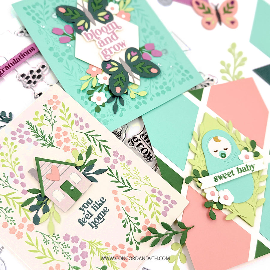 A styled flat-lay of handmade greeting cards featuring botanical and baby-themed designs. The cards include layered green and pink butterflies, a house with a heart, a baby bird, and floral patterns in pastel colors on a white background with small crafting tools.