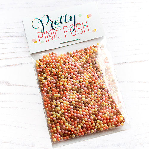A clear plastic bag filled with many small round craft beads in assorted pastel and warm colors, attached to a white paper header card that reads 'Pretty Pink Posh'. The bag is photographed on a light whitewashed wood surface. Pretty Pink Posh Blush Harvest Shaker Beads