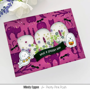 A handmade Halloween greeting card with a pink and purple cat-patterned background, large white 'BOO' word decorated with green leaves, a black ribbon reading 'HAVE A SPOOKY DAY', and cute ghost stickers. The card is photographed on a white surface with flowers in the corner and a watermark at the bottom.