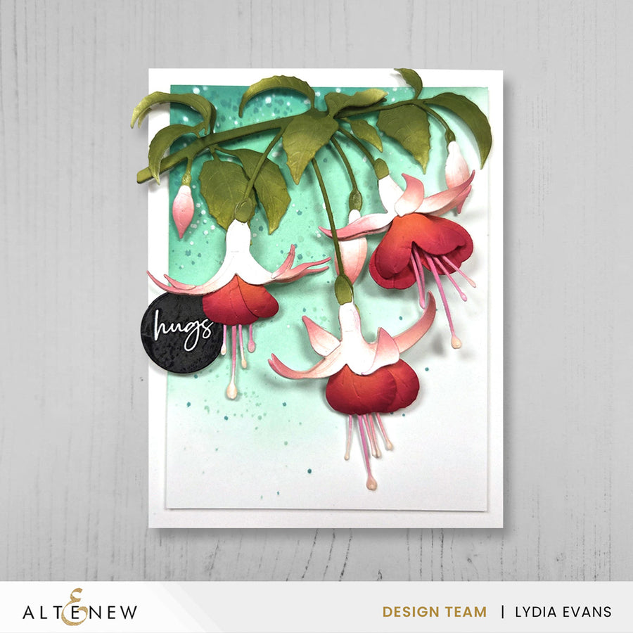 A handmade greeting card featuring layered paper fuchsia flowers in red, pink, and white with green leaves, set against a light teal watercolor background. The card is displayed on a light gray wood-textured background with a white banner at the bottom containing the brand 'ALTENEW' and designer 'LYDIA EVANS' text.