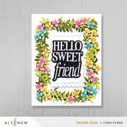 A handmade greeting card featuring the phrase 'HELLO SWEET friend' in black and white lettering, surrounded by a colorful floral border of pink, yellow, and blue flowers with green leaves, displayed on a light gray wood background.