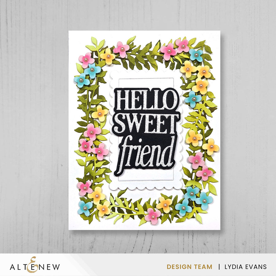 A handmade greeting card featuring the phrase 'HELLO SWEET friend' in black and white lettering, surrounded by a colorful floral border of pink, yellow, and blue flowers with green leaves, displayed on a light gray wood background.