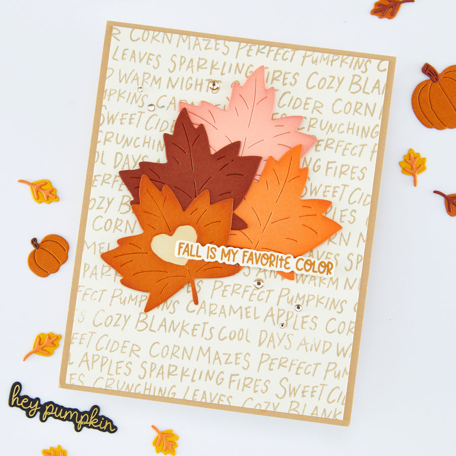 A handmade autumn-themed greeting card featuring layered die-cut maple leaves in shades of orange, brown, and pink, with a small heart-shaped tag that reads 'FALL IS MY FAVORITE COLOR'. The card is displayed on a white background decorated with small pumpkins and scattered fall leaves.