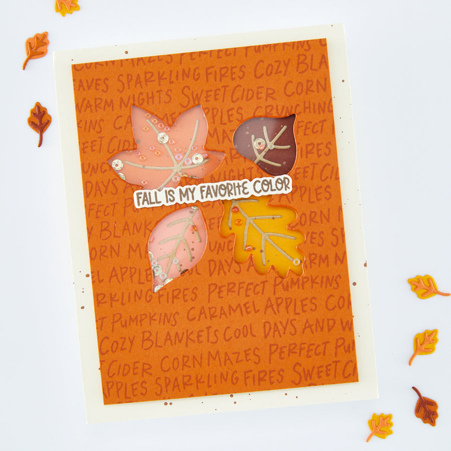 A handmade fall-themed card featuring layered orange and brown leaves, acorns, and a banner that reads 'FALL IS MY FAVORITE COLOR' on a patterned orange background, photographed on a white surface with scattered autumn leaves around it.