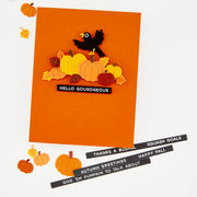 An orange handmade greeting card featuring a layered autumn design with pumpkins, leaves, a black bird, and the text 'HELLO GOURDGEOUS'. Several smaller autumn-themed paper cutouts are scattered around the card on a white background, with additional black text strips reading autumn greetings lying at the bottom right. Hero Arts Autumn Greetings Clear Stamp Set
