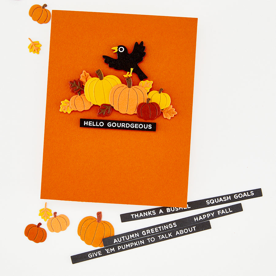 An orange handmade greeting card featuring a layered autumn design with pumpkins, leaves, a black bird, and the text 'HELLO GOURDGEOUS'. Several smaller autumn-themed paper cutouts are scattered around the card on a white background, with additional black text strips reading autumn greetings lying at the bottom right. Hero Arts Autumn Greetings Clear Stamp Set