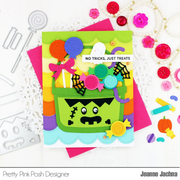 A handmade Halloween greeting card featuring a green basket filled with colorful craft elements, shown on top of pink cards with decorative craft tools and buttons around it. The card reads 'NO TRICKS, JUST TREATS' and is branded Pretty Pink Posh Designer and Jeanne Jachma.