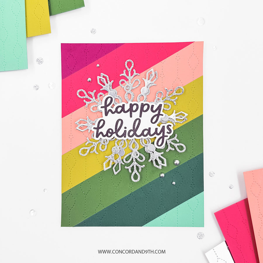 A handmade holiday greeting card featuring diagonal stripes in magenta, pink, peach, yellow, green, and teal, with a silver glittery snowflake and the words 'happy holidays' in dark gray script. The card is displayed on a white background with scattered decorative gems and partial colorful paper stacks at the edges.