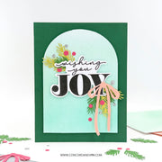 A handmade Christmas greeting card with a dark green border, a light aqua arched center panel, and layered holiday embellishments including pine branches, berries, and a peach ribbon. The card reads 'wishing you JOY' in black and white lettering.
