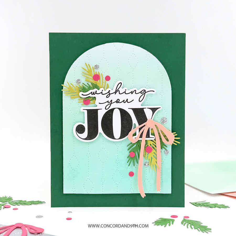 A handmade Christmas greeting card with a dark green border, a light aqua arched center panel, and layered holiday embellishments including pine branches, berries, and a peach ribbon. The card reads 'wishing you JOY' in black and white lettering.