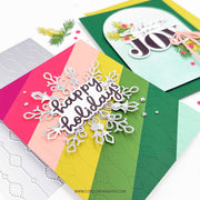 A close-up of a handmade holiday greeting card with diagonal rainbow stripes in pink, peach, yellow, green, and mint, featuring a silver glittery snowflake and the words 'happy holidays.' Another card in the background has 'wishing you joy' in layered die-cut letters.
