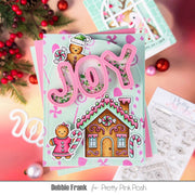 A handmade Christmas card featuring pink 3D 'JOY' lettering with glitter, a gingerbread house, and gingerbread people, styled with holiday greenery and ornaments on a pink background. The text 'Debbie Frank for Pretty Pink Posh' is visible at the bottom.