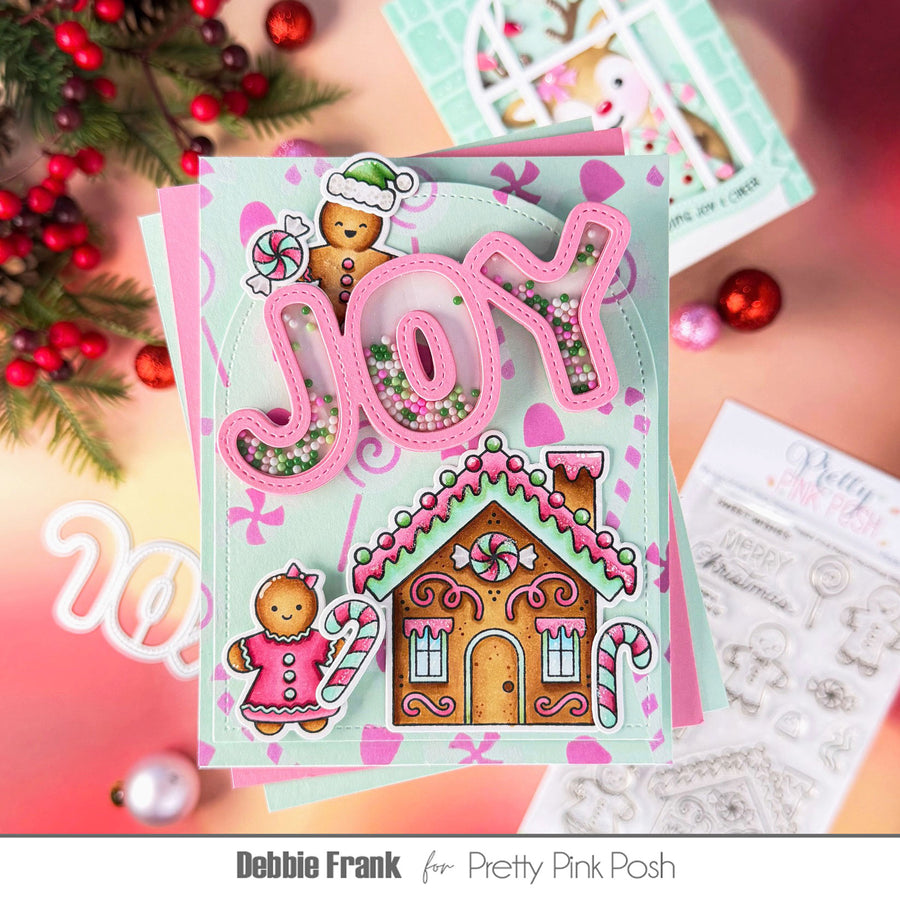 A handmade Christmas card featuring pink 3D 'JOY' lettering with glitter, a gingerbread house, and gingerbread people, styled with holiday greenery and ornaments on a pink background. The text 'Debbie Frank for Pretty Pink Posh' is visible at the bottom.