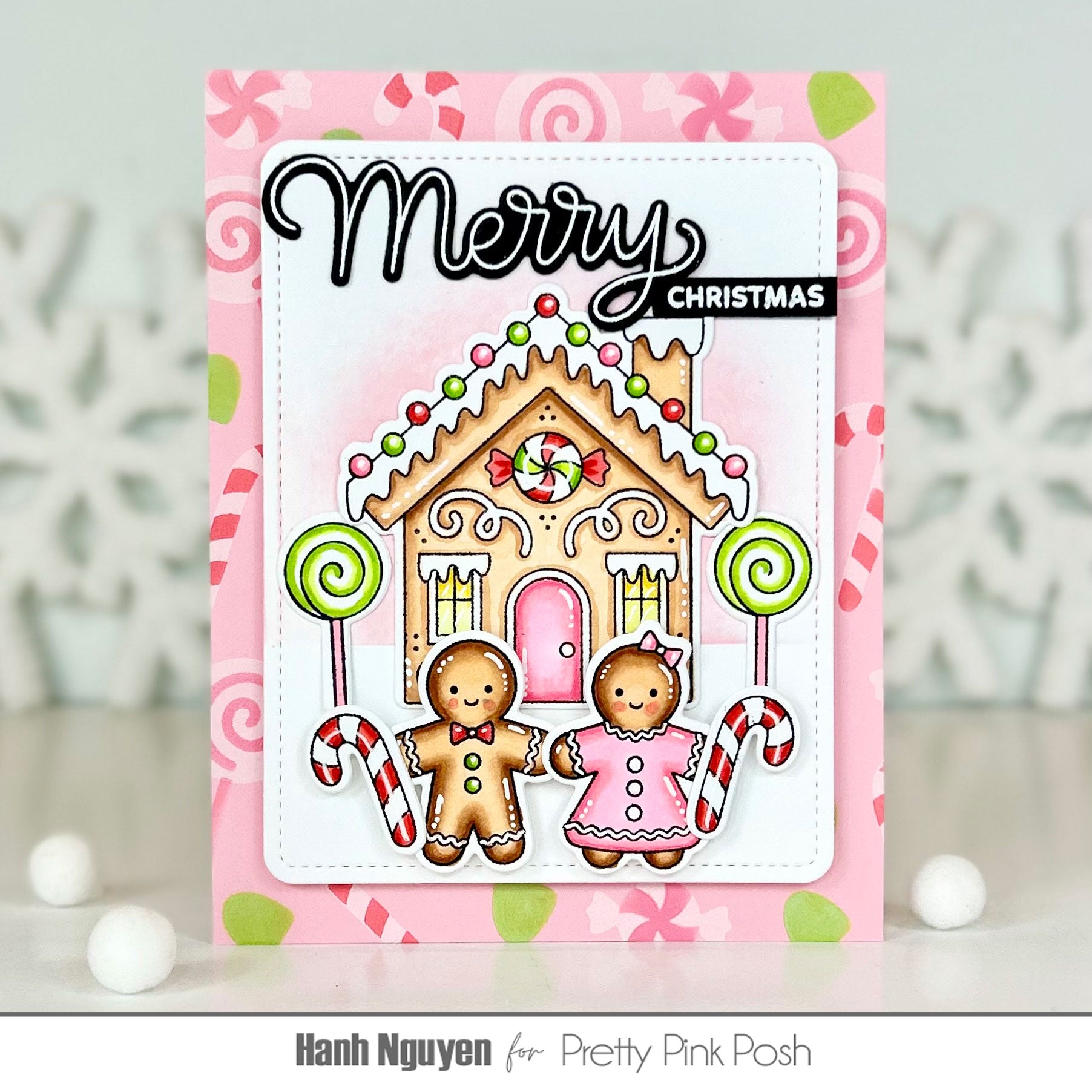 A handmade Christmas greeting card featuring a gingerbread house, two gingerbread people, candy canes, and the words 'Merry Christmas' on a pink patterned background. The card is displayed upright on a light surface with white decorative snowflakes in the background.