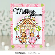 A handmade Christmas greeting card featuring a gingerbread house, two gingerbread people, candy canes, and the words 'Merry Christmas' on a pink patterned background. The card is displayed upright on a light surface with white decorative snowflakes in the background.