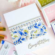 A close-up flat-lay of a handmade greeting card with a white embossed base, a decorative border of blue and yellow flowers, and the word 'Congratulations' in metallic script. The card is styled on a pink surface with craft supplies and a visible Rachael Brand stamp box nearby.