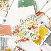 A beautifully arranged flat-lay of handmade greeting cards and stamping supplies from the brand Paperwhisk. The central focus is a cream-colored card featuring a colorful floral pattern in red, orange, yellow, and green, with metallic 'Congratulations' text.