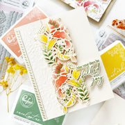 A layered die-cut card featuring a floral vine design in orange, yellow, green, and white, with the phrase 'May your days be filled with joy' in green script. The card is styled on top of other crafting supplies, including embossed paper, dye ink pads, and stamps, with small yellow flowers as props.