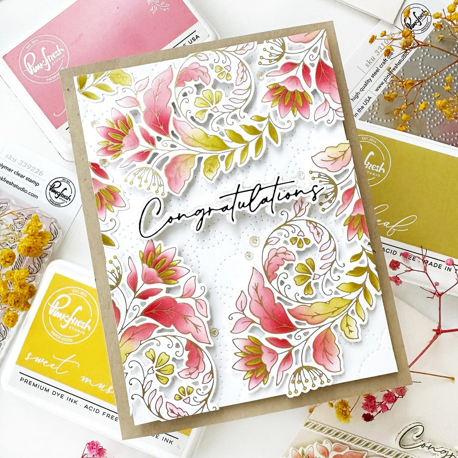 A handmade congratulations card featuring layered floral die-cut designs in pink, yellow, and green on a white background with a kraft paper border. The card is styled on a flat-lay surface surrounded by crafting supplies, including Ruffwafers ink pads and dried flowers.