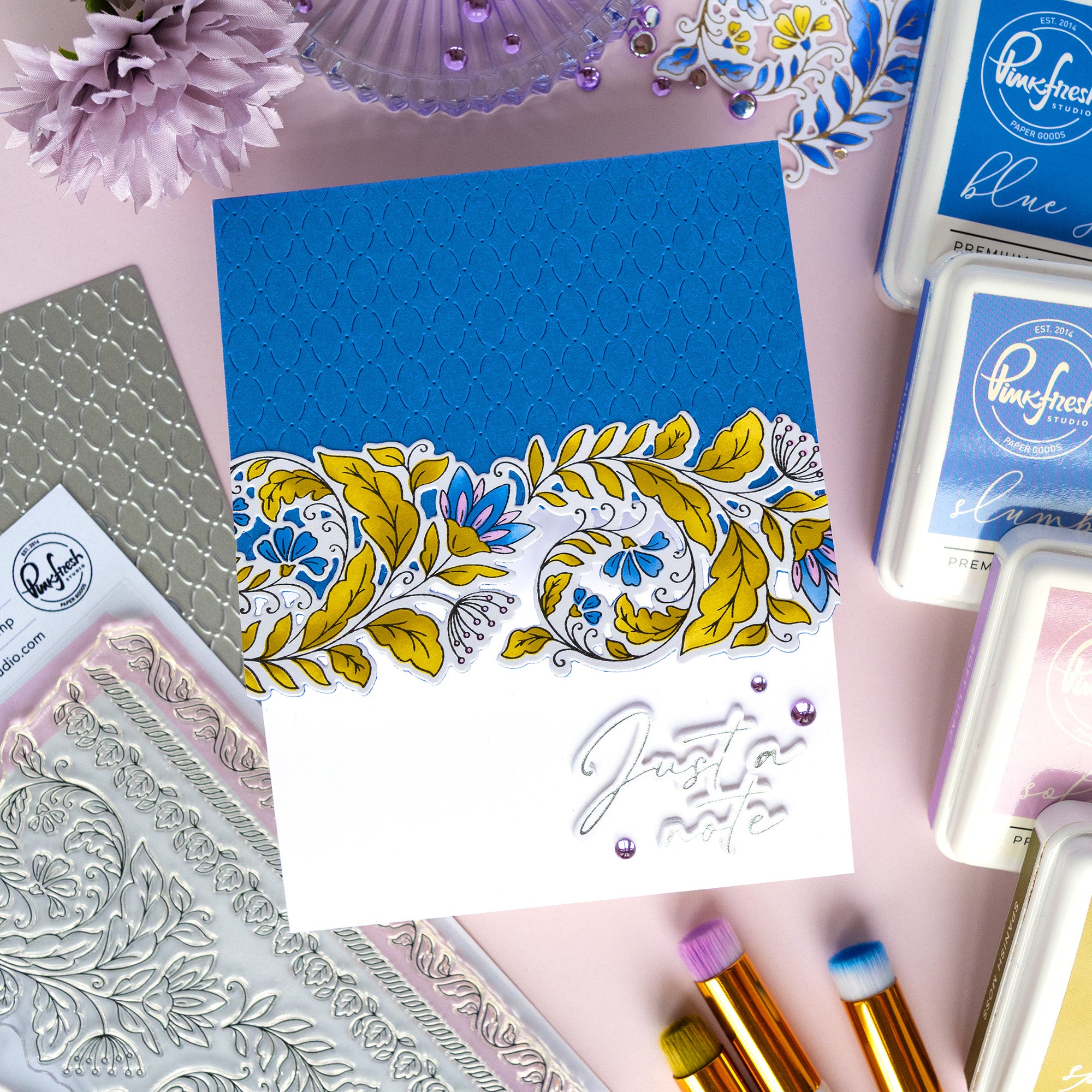 A handmade greeting card with a blue and white design, featuring a yellow and blue floral border and the phrase 'Just a smile' in silver foil script. The card is styled in a flat-lay arrangement with crafting tools, stamps, and ink pads on a pink background.