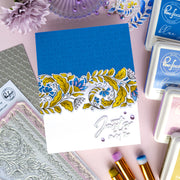A handmade greeting card with a blue and white design, featuring a yellow and blue floral border and the phrase 'Just a smile' in silver foil script. The card is styled in a flat-lay arrangement with crafting tools, stamps, and ink pads on a pink background.