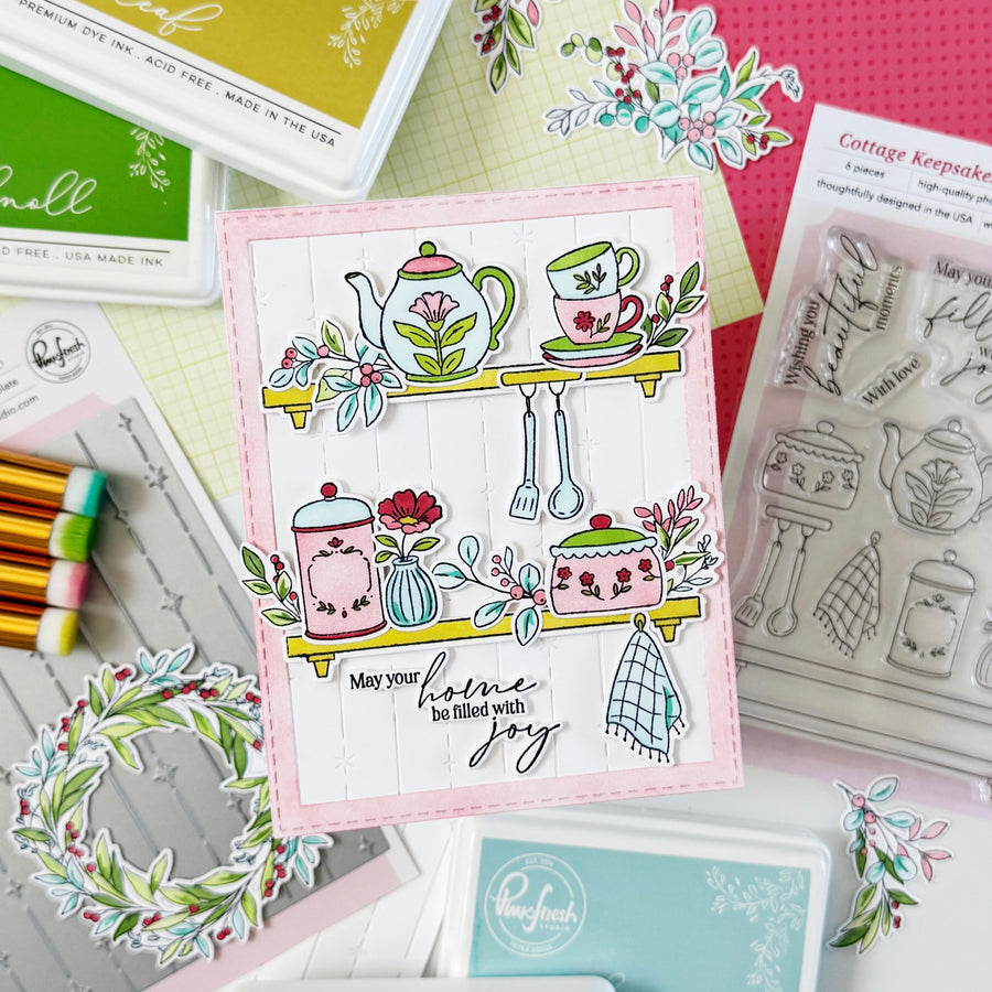A handmade greeting card featuring a whimsical kitchen shelf illustration with a teapot, teacup, flowers, jars, and the phrase 'May your home be filled with joy,' styled in a flat-lay with crafting supplies and stamps.