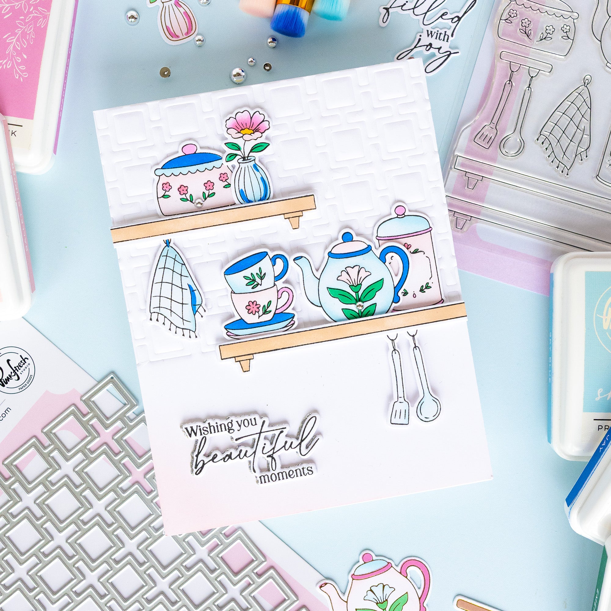 A handmade layered card featuring illustrated kitchen items like a teapot, teacup, flowers, and utensils on wooden shelves, with the stamped phrase 'Wishing you beautiful moments' at the bottom, styled on a pastel craft background with other cards and stamping supplies.