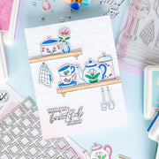 A handmade layered card featuring illustrated kitchen items like a teapot, teacup, flowers, and utensils on wooden shelves, with the stamped phrase 'Wishing you beautiful moments' at the bottom, styled on a pastel craft background with other cards and stamping supplies.