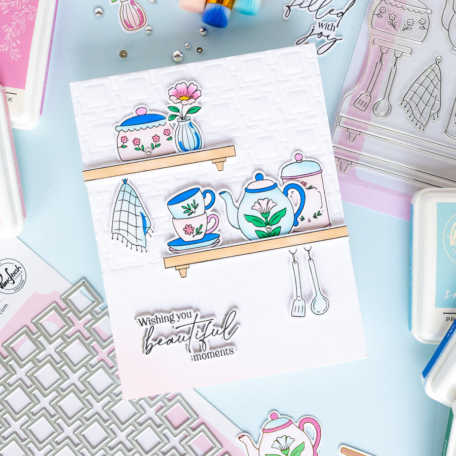 A handmade layered card featuring illustrated kitchen items like a teapot, teacup, flowers, and utensils on wooden shelves, with the stamped phrase 'Wishing you beautiful moments' at the bottom, styled on a pastel craft background with other cards and stamping supplies.