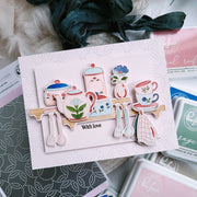 A handmade layered card featuring whimsical tea-time illustrations including a teapot, teacup, sugar bowl, flowers, and utensils on a pale pink textured background, styled with dye ink pads and fabric in a flat-lay arrangement.