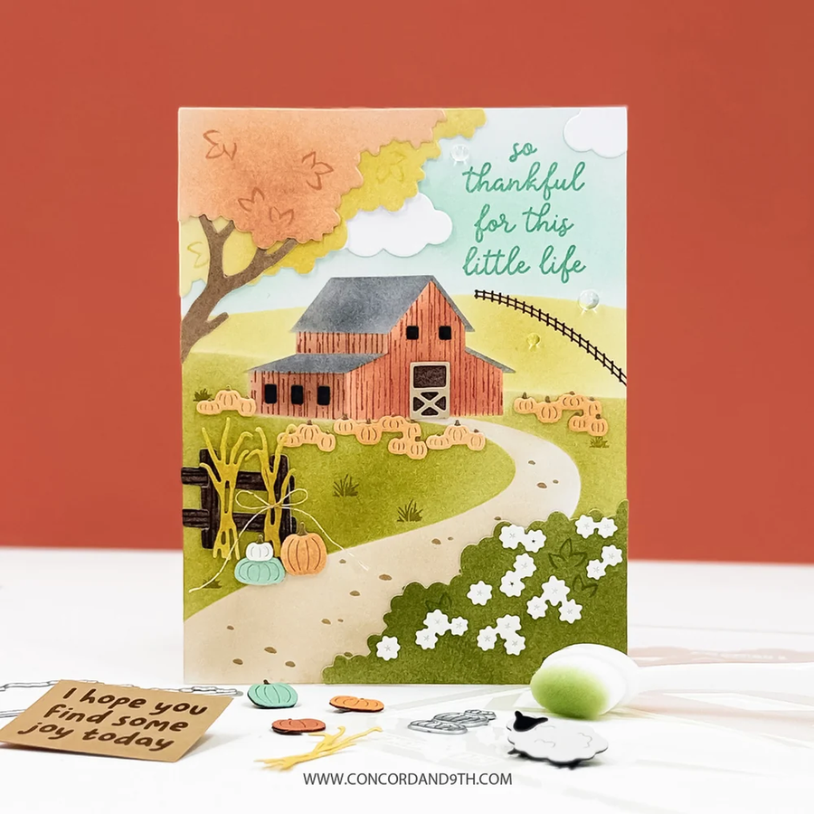 A handmade layered card featuring a rustic autumn farm scene with a red barn, pumpkins, flowers, and the text 'so thankful for this little life.' The card is displayed upright on a white surface with small crafting props and a cardstock tag in front, against a warm orange background.