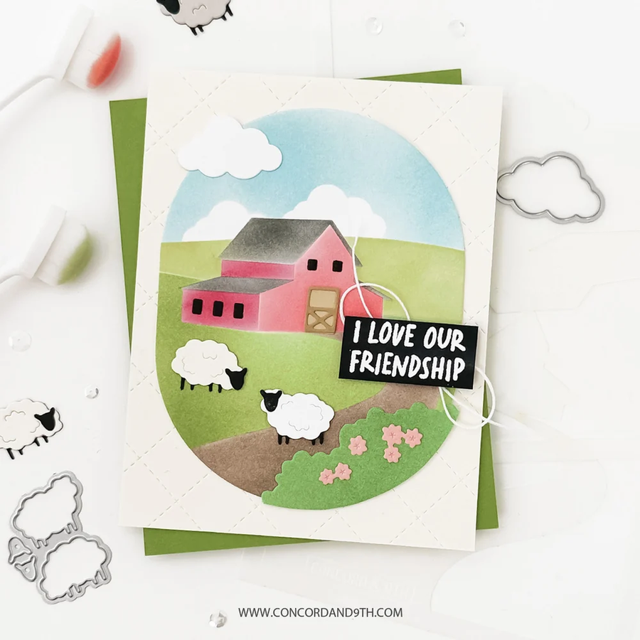 A handmade layered card featuring a colorful farm scene with a pink barn, green hills, sheep, clouds, and the text 'I LOVE OUR FRIENDSHIP'. The card is displayed on a white surface with crafting tools and metal dies around it.