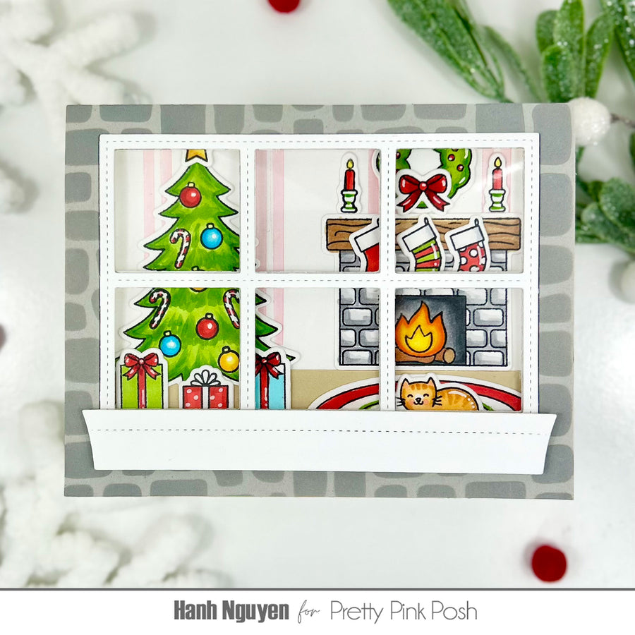 A handmade Christmas-themed window card with six panes showing holiday scenes: a Christmas tree, candles, stockings, a fireplace, candy canes, and a cat. The card is styled on a white surface with festive greenery and red ornaments around it.