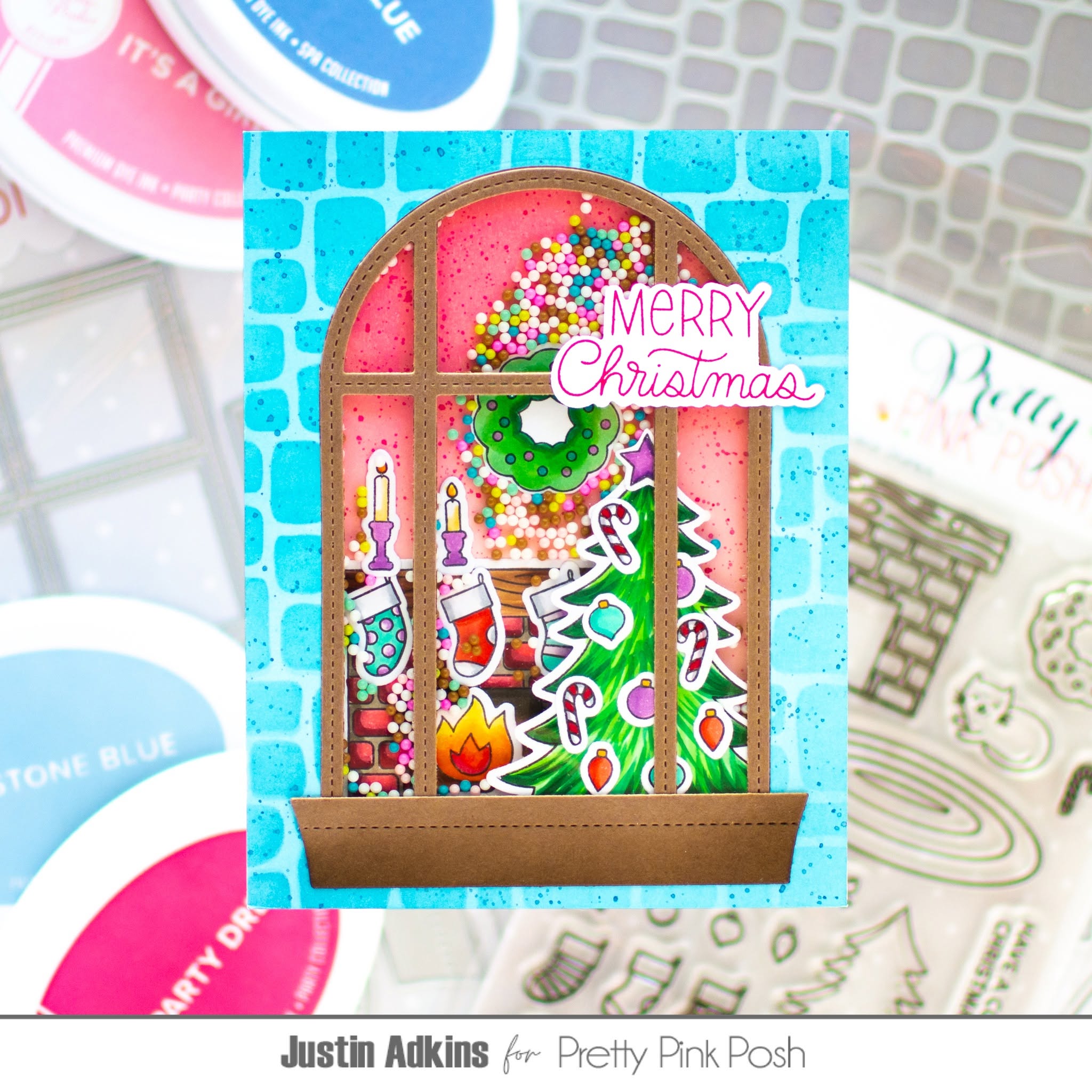A handmade Christmas window card featuring a colorful festive scene with candy canes, ornaments, candles, and a green wreath under a pink sky, with 'Merry Christmas' written in pink script. The card is displayed on a light blue brick-patterned background with craft supplies and stamps visible around it.