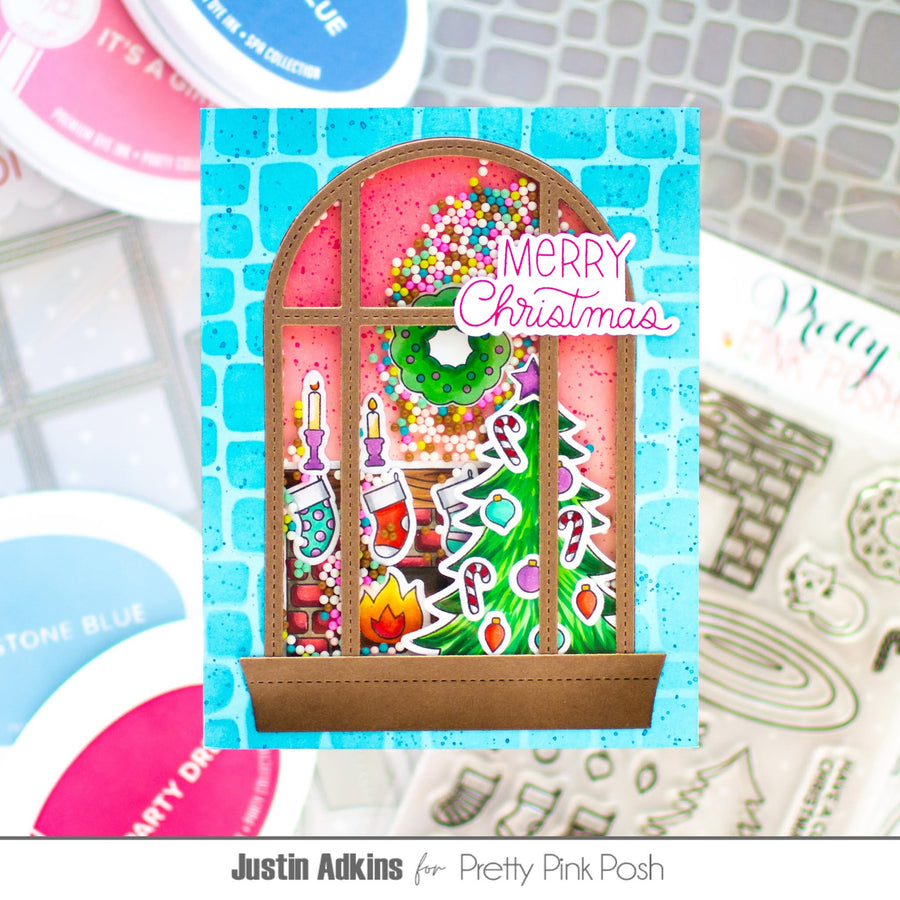 A handmade Christmas window card featuring a colorful festive scene with candy canes, ornaments, candles, and a green wreath under a pink sky, with 'Merry Christmas' written in pink script. The card is displayed on a light blue brick-patterned background with craft supplies and stamps visible around it.