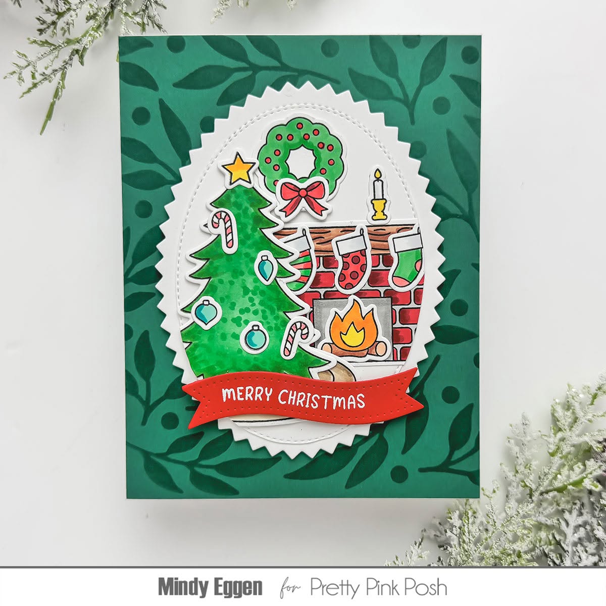 A handmade Christmas greeting card featuring a green leafy patterned background, a scalloped white oval layer with a Christmas tree, wreath, stockings, fireplace, and candle illustration, and a red banner reading “Merry Christmas.” The card is photographed on a white surface with decorative foliage at the corners.