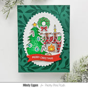 A handmade Christmas greeting card featuring a green leafy patterned background, a scalloped white oval layer with a Christmas tree, wreath, stockings, fireplace, and candle illustration, and a red banner reading “Merry Christmas.” The card is photographed on a white surface with decorative foliage at the corners.