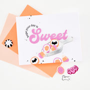 A white greeting card with a pink 'Sweet' script and cookie illustrations, styled on top of peach and orange envelopes with small decorative cookies on a white background.
