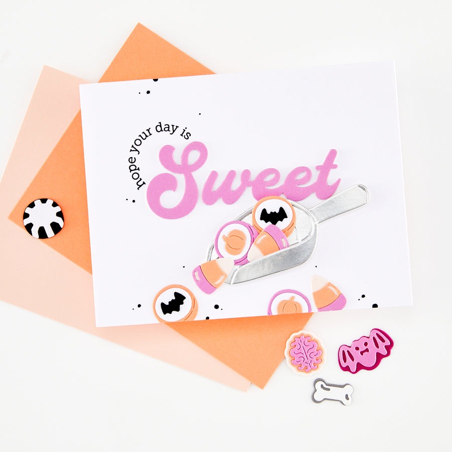 A white greeting card with a pink 'Sweet' script and cookie illustrations, styled on top of peach and orange envelopes with small decorative cookies on a white background.