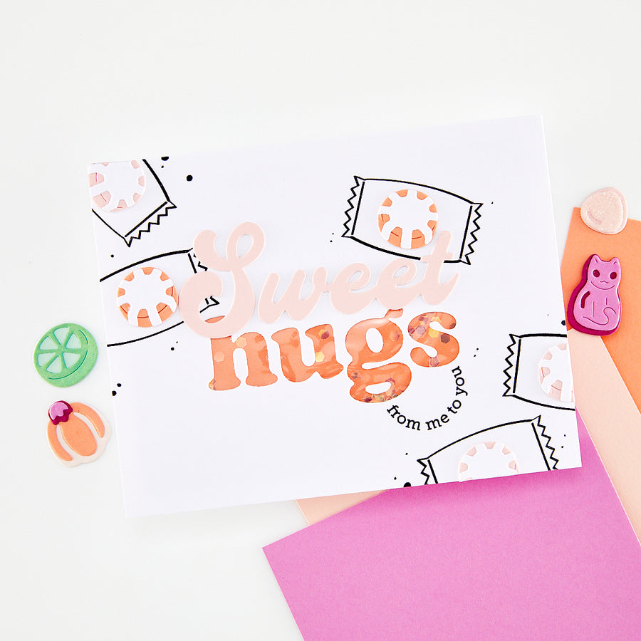 A white handmade greeting card with the words 'Sweet hugs from me to you' in peach and orange lettering, surrounded by pastel paper and small decorative items on a white background.
