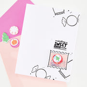 A white handmade greeting card with black candy illustrations and the text 'sending SWEET holiday wishes,' styled on pink and peach paper with small decorative craft pieces. The card is the main product shown in a clean flat-lay setup.