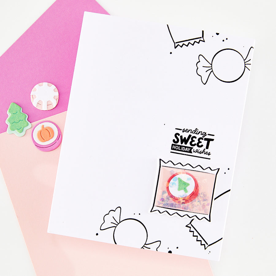 A white handmade greeting card with black candy illustrations and the text 'sending SWEET holiday wishes,' styled on pink and peach paper with small decorative craft pieces. The card is the main product shown in a clean flat-lay setup.