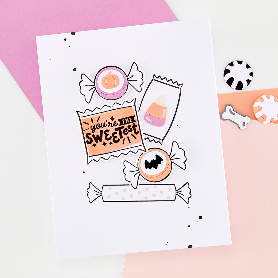 A white handmade greeting card featuring cute Halloween candy illustrations, styled on a pink and white background with small matching candy-shaped accessories nearby.