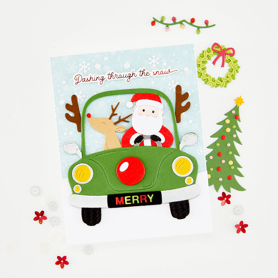 A festive handmade Christmas card featuring Santa Claus driving a green car with reindeer antlers, surrounded by holiday decorations including a wreath, Christmas tree, garland, and scattered red flowers on a white background. Using Hero Arts Beep Beep Die Set