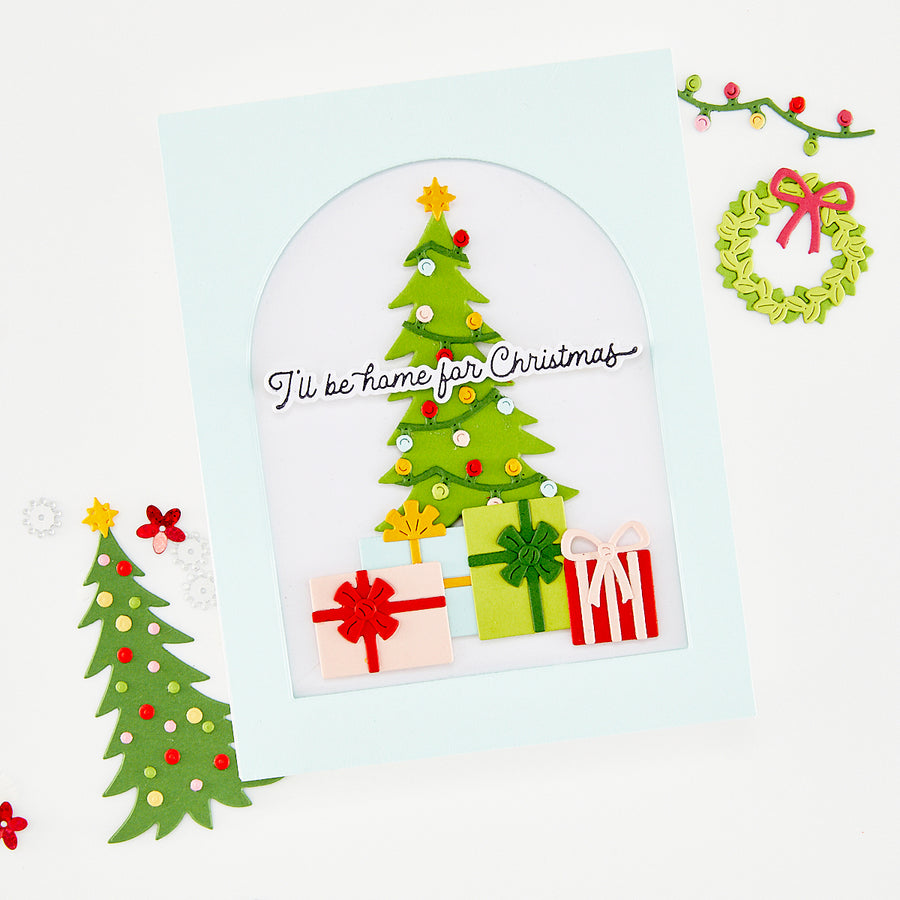 A handmade layered paper Christmas card with a light blue border, featuring a green paper Christmas tree with colorful ornaments, wrapped gift boxes, and the text 'I'll be home for Christmas'. The card is styled on a white background with decorative paper holiday elements. Using Hero Arts Beep Beep Holiday Extras Die Set