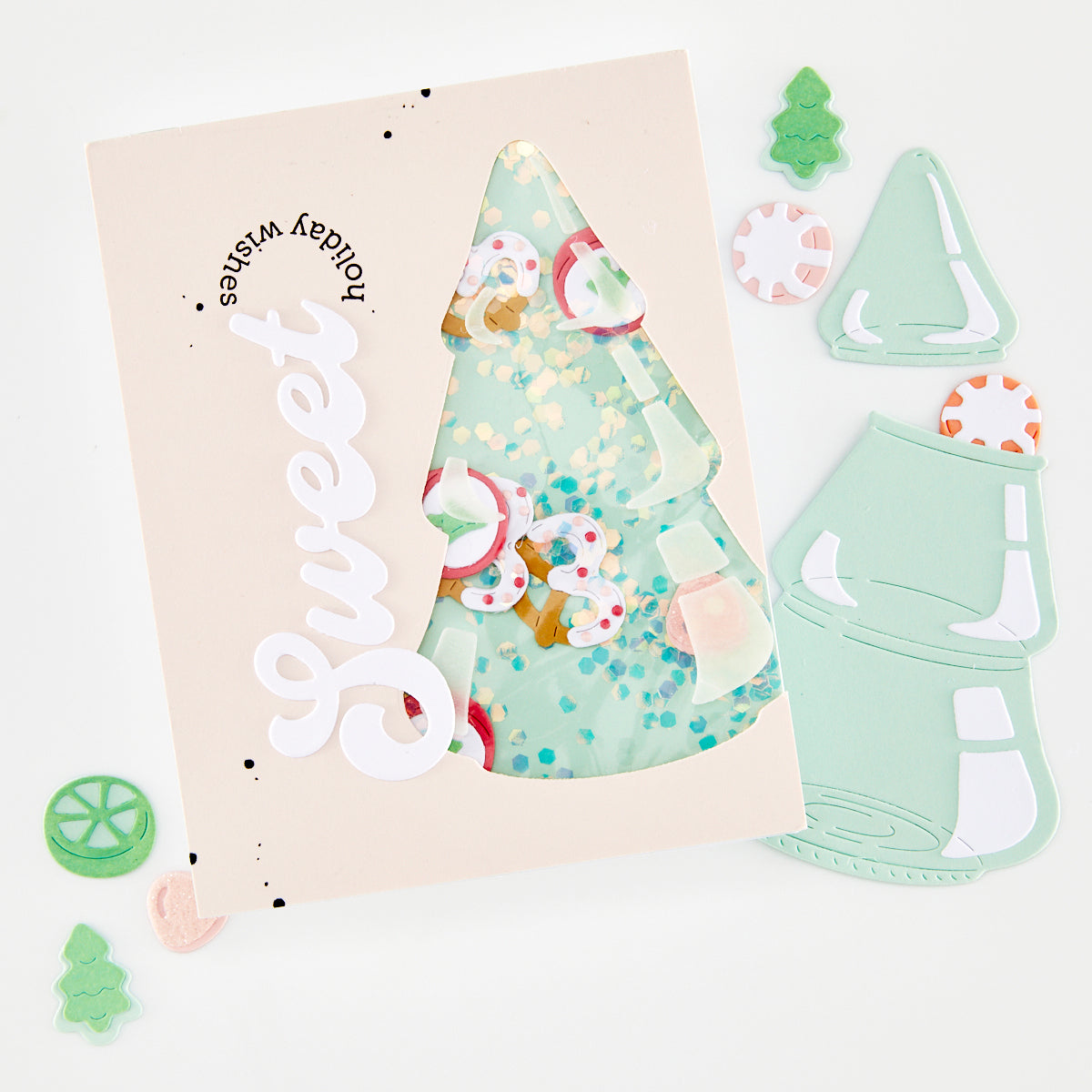 A flat-lay of a handmade greeting card with a die-cut white 'Sweet' script and a window showing a mint green Christmas tree filled with colorful confetti and candy decorations. Surrounding the card are matching mint green die-cut embellishments, including a Christmas tree, a candy cane, and a gingerbread man, on a clean white background.