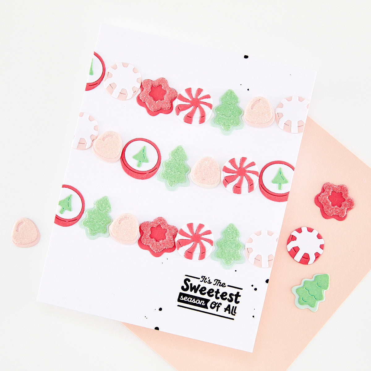 A white greeting card with a festive Christmas garland design made of red, pink, and green die-cut stickers, displayed on a peach envelope with scattered matching stickers on a white background.