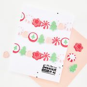 A white greeting card with a festive Christmas garland design made of red, pink, and green die-cut stickers, displayed on a peach envelope with scattered matching stickers on a white background.